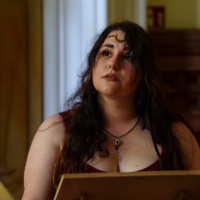 Woman with dark wavy hair and a moon forehead jewelry sits at a wooden drawing board, gazing upward in thought, with an ink bottle nearby.