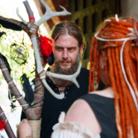 Bearded man in a black lace-up shirt faces a person with orange dreadlocks, both in medieval costumes with antlered staff props in a doorway setting.