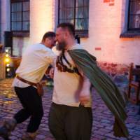 Two men in medieval costumes dancing or sparring in a cobblestone courtyard under string lights, one with a green cape.