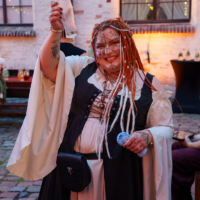 Person in medieval fantasy costume raises a glass in a courtyard at dusk, smiling with decorative face paint and orange-dreadlock wig.