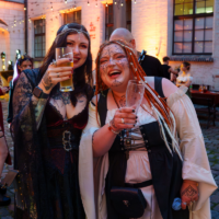 Two women in medieval/fantasy costumes raise glasses at an outdoor party under string lights.