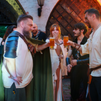 Group of people in medieval costumes raise beer mugs in a toast outdoors near a stone archway, with others watching and smiling.