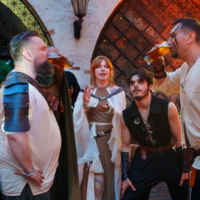 Group of adults in medieval costumes sharing a beer toast outdoors near arched stone windows at a Renaissance fair.