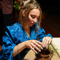 Woman in a blue satin dress pours from a bottle into a copper pot on a patterned table, performing a ritual or ceremony.