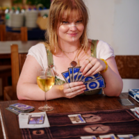 Woman smiling and dealing cards at a wooden table, fanning blue-backed cards with a glass of white wine nearby.