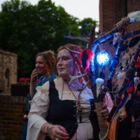 Person in a fantasy costume with antler branches and a glowing blue-lit staff at an outdoor festival, face painted with designs.