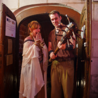 Two costumed adults stand in a dungeon doorway; the man holds a spiked wooden mace while the woman, in a flowing cream dress, covers her mouth in surprise.