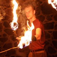 Fire performer in a red shirt spins flaming torches inside a stone-walled room.