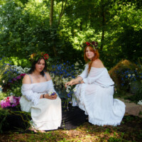 Two women in white dresses with flower crowns sit among wildflowers in a sunlit garden setting.