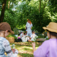 Group of people in a sunlit forest clearing; two hat-wearing participants in the foreground offer flowers to three women in white dresses with flower crowns.