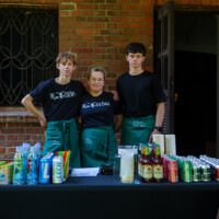 Three volunteers stand behind a beverage table outdoors, posing in black shirts and teal aprons in front of a brick wall with a window and door behind them.
