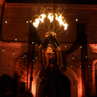 Two fire dancers raise a burning hoop above their heads during an outdoor evening performance under string lights.