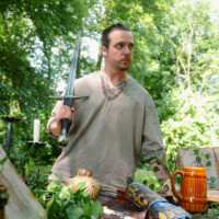 Man in a beige tunic holds a long sword at a table of medieval props during an outdoor fair or market setting.