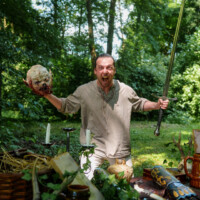 Man in a beige tunic in a forest, raising a long sword in one hand and a prop severed head in the other, with medieval props on a table nearby.