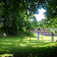 Two people in white clothing practicing archery in a sunlit park with rope barriers and lush trees, targets to the left and a brick wall in the background