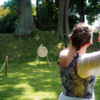 A person outdoors in a park aiming a bow at a round straw archery target on a stand, with trees and sunlight in the background.