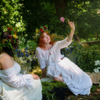 Two women in white dresses wearing flower crowns sit among a vibrant garden bouquet, crafting a floral display.