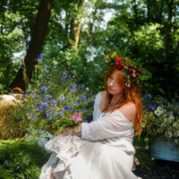 Red-haired woman in a flowing white dress with a flower crown sits among wildflowers in a sunlit forest clearing.