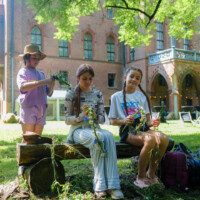 Three girls sit on a log crafting flower garlands in a sunny campus garden with a brick building in the background.