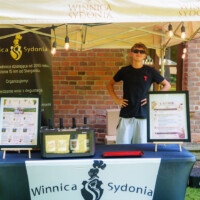 Young man in sunglasses behind a Winnica Sydonia wine stall under a canopy with string lights.