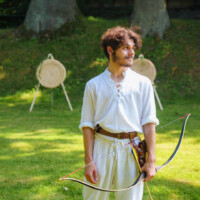 Young man in white medieval-style shirt and belt holds a bow outdoors, with archery targets in the background on a sunny lawn.