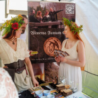 Two women in white period dresses with flower crowns chat at a fantasy booth, exchanging cards over a table of brochures and small items.