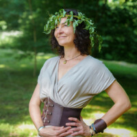 Woman outdoors wearing a gray wrap dress and a wide leather belt, with a laurel wreath on her head, smiling confidently.