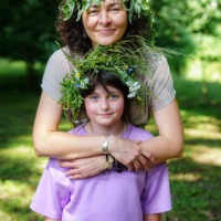 Mother embraces daughter in a park, both wearing leafy flower crowns made of greenery and wildflowers.