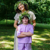 Smiling woman and girl wearing leafy flower crowns stand in a sunlit park.