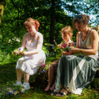 Three people—two adults and a child—sit on a log outdoors, weaving wildflowers into flower garlands in a sunlit park.”] ,
