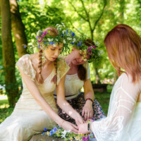 Three women in a sunlit forest weave colorful wildflowers into flower crowns together around a wooden bench.