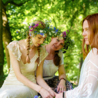 Three women wearing flower crowns craft a floral garland together in a sunlit forest clearing.