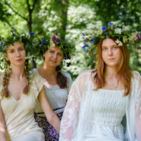 Three women in white dresses wearing large flower crowns pose in a sunlit, leafy park.