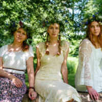Three women in white dresses with flower crowns sit on a log in a sunlit park.