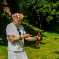 Older woman with a colorful flower crown drawing a bow and aiming an arrow outdoors in a sunny park.