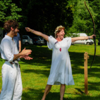 Woman in a white dress with a flower crown stretches her arms while holding a bow, smiling in an outdoor grassy area as a man in white claps nearby.