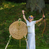 Woman in white with a flower crown and sunglasses raises her fist while holding a bow beside a large woven circular archery target in a grassy outdoor area.