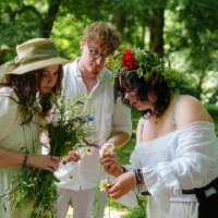 Three friends in a sunlit park wearing white, gathering wildflowers and arranging a flower crown on one person’s head.