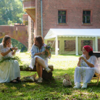 Three women in white dresses weave flowers into garlands on a sunny lawn next to a brick building and white tent teepee in the background.