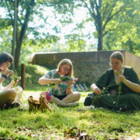 Three women sit on the grass weaving wildflowers into garlands in a sunny park setting.