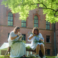 Two women in white dresses sit on a wooden bench weaving wildflowers, outdoors under leafy trees with a brick building in the background.