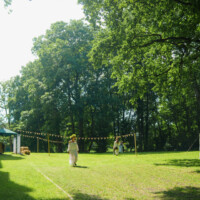 Sunlit grassy park with decorative bunting and a few people walking toward a gathering among tall green trees.