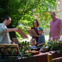 Man showing a vintage book to a family at an outdoor antique stall with various objects on a table under trees.