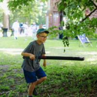 Boy with glasses and a cap runs with a black foam bat in a park under green leaves.