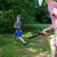 Boy in a gray shirt and blue shorts dodges a foam sword while an adult holds a foam baton in a sunlit park.
