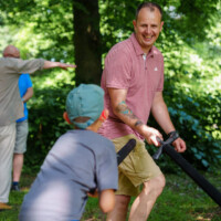 Man in a pink polo shirt smiles while guiding a foam baton in a park as a child approaches; others are in the background among trees and greenery.