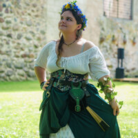 Woman in a Renaissance-style dress with a floral belt and green skirt, wearing a blue-yellow flower crown, outdoors by a stone wall.