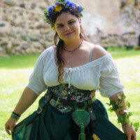 Woman in a floral crown and medieval-style outfit outdoors near an old stone wall, posing for the camera.
