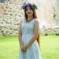 Young woman in a white dress with a colorful flower crown standing on green grass with a stone wall behind her.