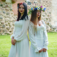 Two women in white dresses pose outdoors by a stone wall; one wears a flower crown and bright lipstick.